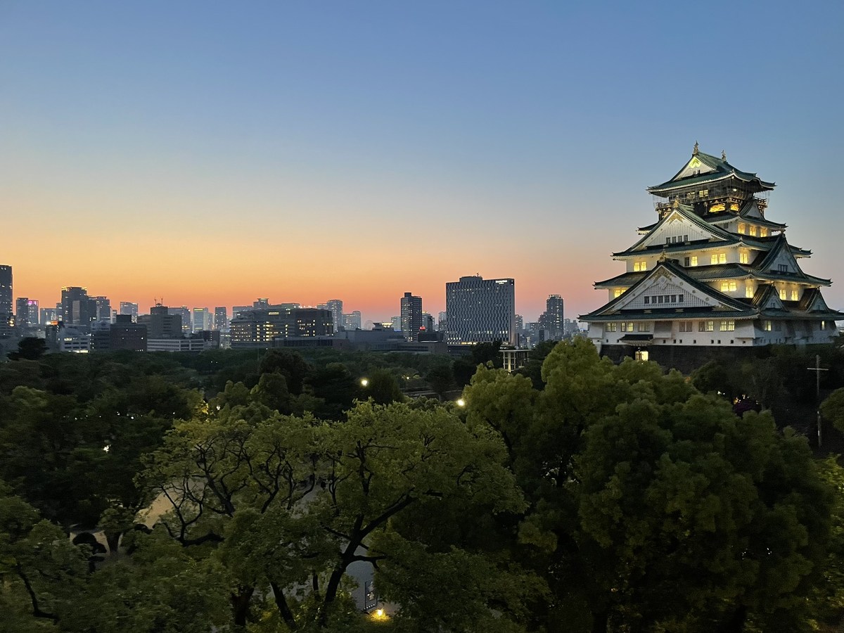 An illuminated Japanese castle above trees and a city skyline at sunset